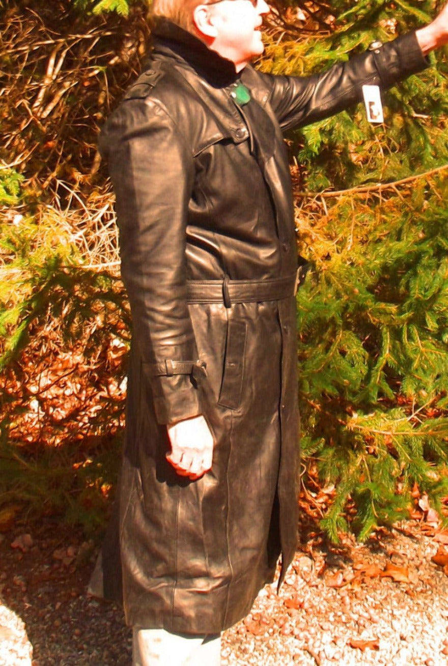 Picture of a model wearing our mens black leather trench coat full length side nt view with green foliage in the background.