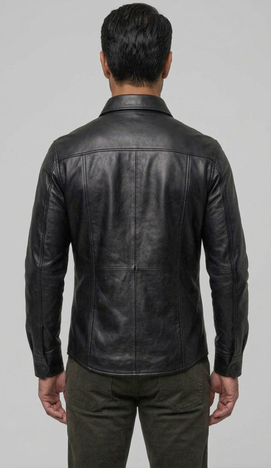 A man viewed from the back wearing our  Black Custom Leather Shirt for Men and dark olive pants against a light gray studio background.
