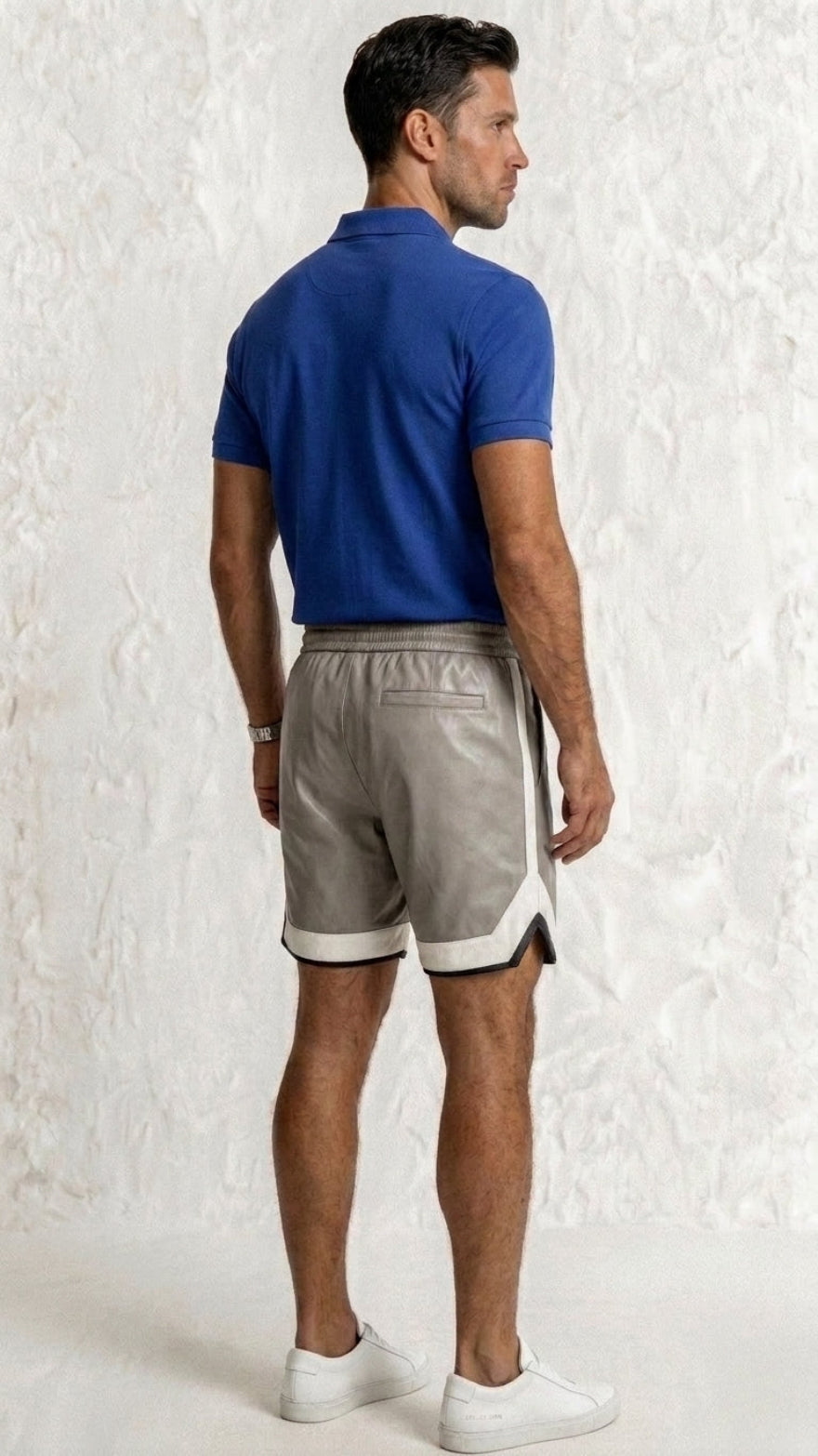 Man wearing a blue polo shirt and gray leather shorts on a textured white background , back view