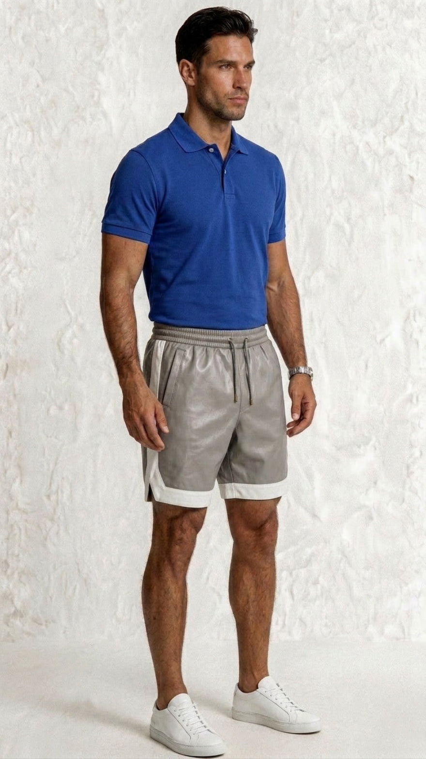 Man wearing a blue polo shirt and  gray leather shorts against a textured white wall side view