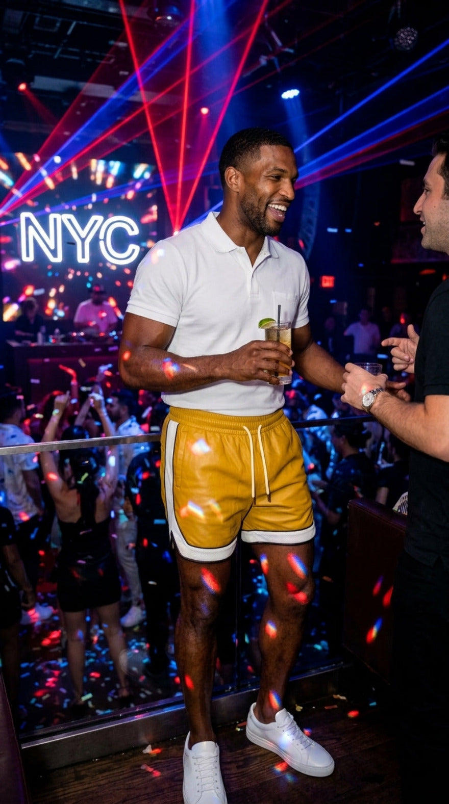 Picture of a man wearing our Yellow Men's Leather Shorts with Contrast Side Stripe and a white polo shirt He has  a drink in his hand, talking in a nightclub.