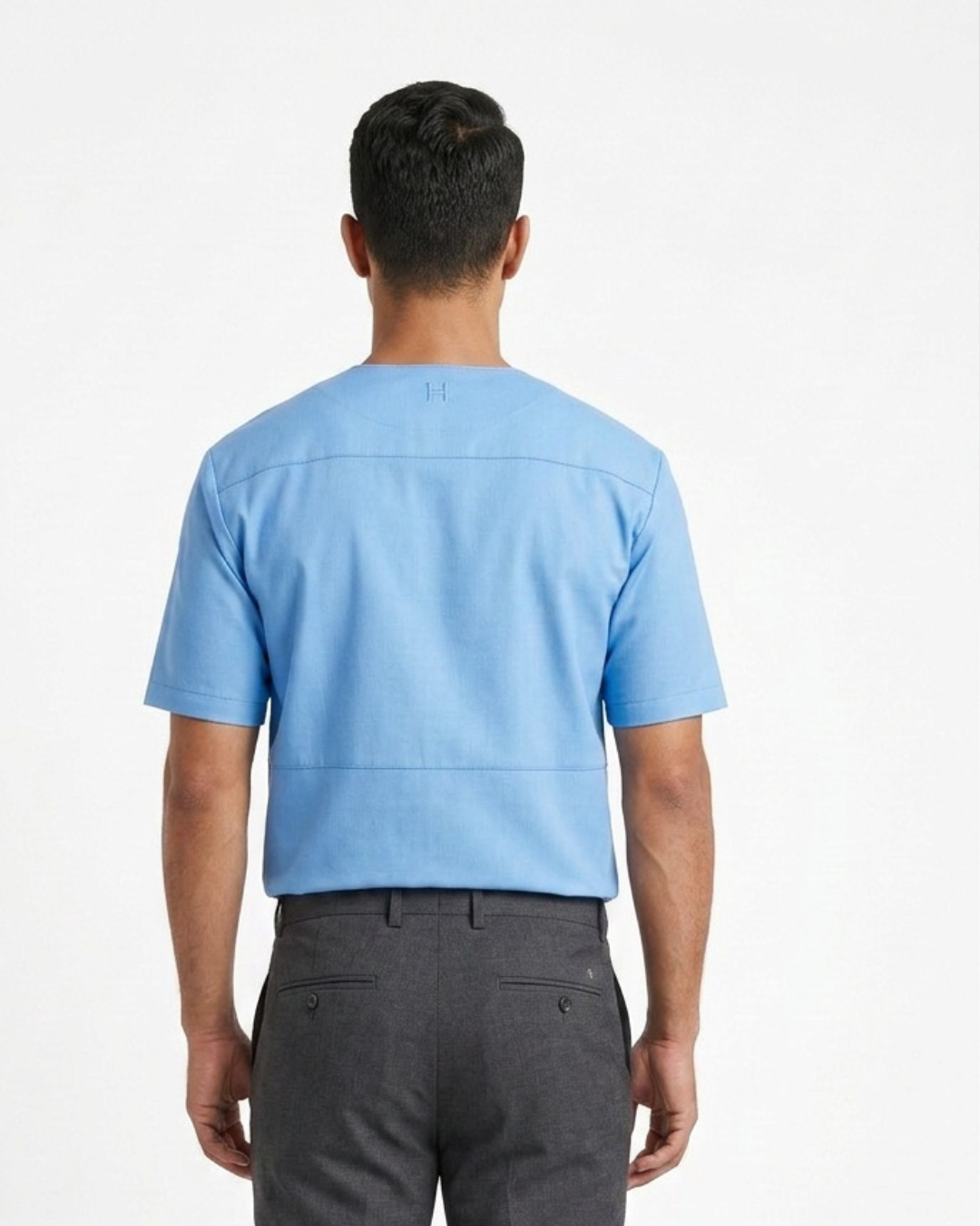 A man viewed from behind wearing a light blue short-sleeve leather tee shirt  and dark gray trousers, shot against a plain white studio background.