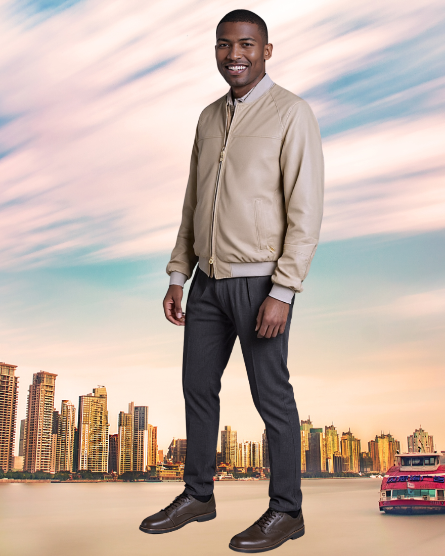 Man in a beige jacket and dark pants standing in front of a city skyline with a colorful sky.
