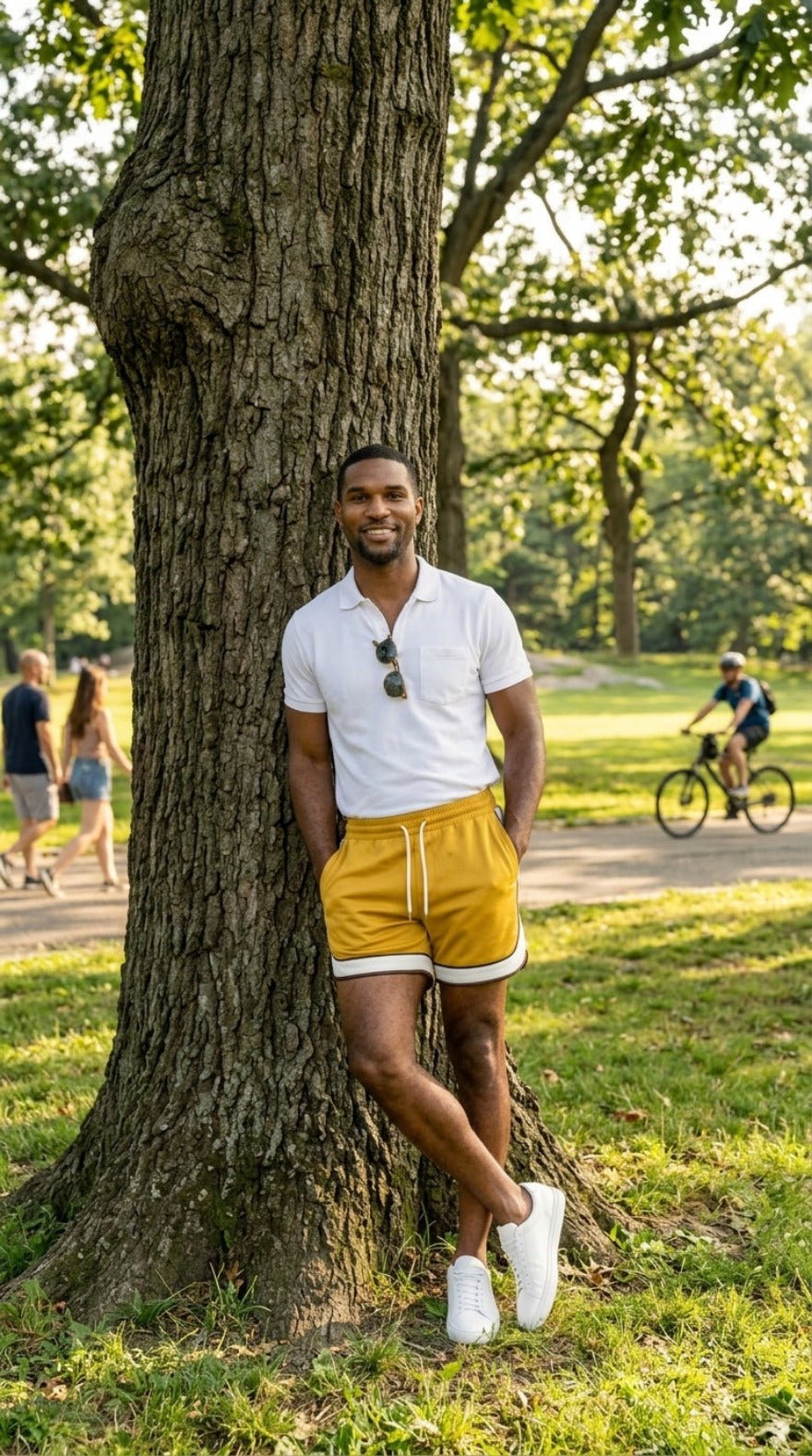 Picture of a man wearing our Yellow Men's Leather Shorts with Contrast Side Stripe and a white polo shirt,  leaning against a tree in  a park.